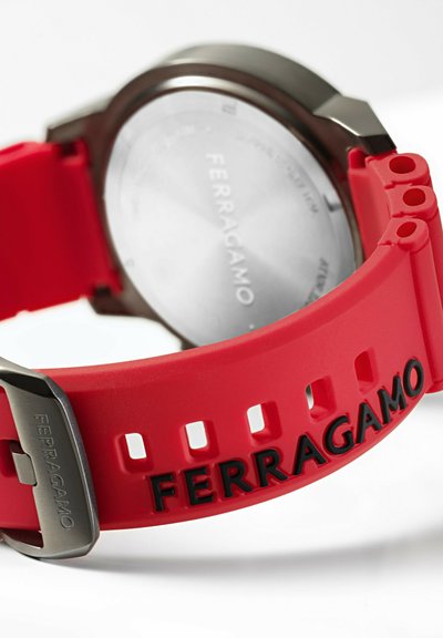 Czerwona silikonowa bransoleta w zegarku z wyciętymi otworami, z metalową kopertą w kolorze srebrnym i grawerem "Ferragamo" na bransolecie, prezentująca minimalistyczny design.