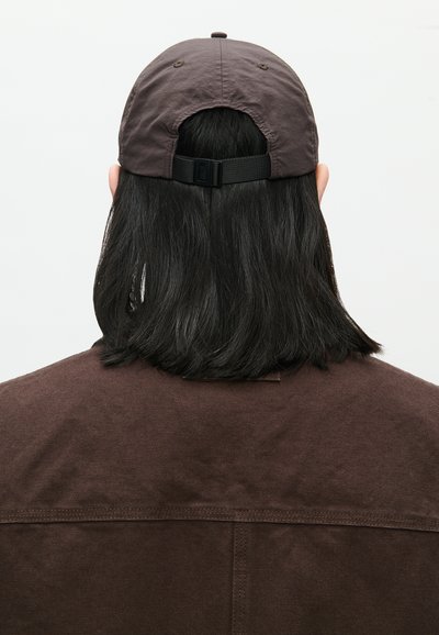 Asmuo su pečių ilgumo juodais plaukais, dėvintis rudą kepurę ir atitinkantį rudą švarką, matomas iš nugaros priešais vientisą foną.