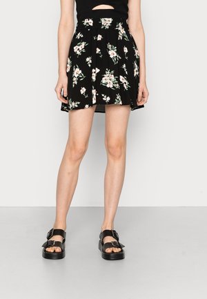 Jupe florale noire avec un design plissé, ornée de fleurs blanches et vertes. Associée à des sandales noires, mettant en avant un détail de boucle chunky.