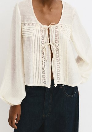 Blouse blanche transparente avec bordure en dentelle, détails plissés, et deux attaches à nouer à l'avant, portée avec un jean en denim foncé.