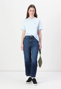 Camiseta de algodón azul claro, jeans rectos de mezclilla azul oscuro, zapatillas negras y un bolso verde. Atuendo simple y casual con un detalle mínimo.