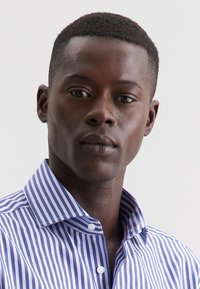 Giovane uomo di colore con capelli corti, che indossa una camicia a righe blu e bianche, su uno sfondo chiaro e uniforme.