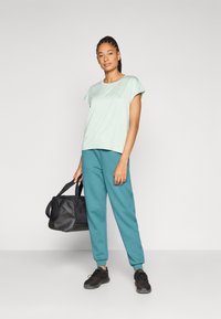 Ανοιχτό πράσινο κοντομάνικο μπλουζάκι με στρογγυλή λαιμόκοψη, συνδυασμένο με πετρόλ παντελόνι τζόγκερ και μαύρα αθλητικά παπούτσια. Το μοντέλο κρατάει μια μαύρη τσάντα duffel.