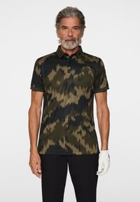 Kortsleeved polo tröja i mörkgrön och svart, med ett zigzag-mönster, knappslå och krage, kombinerad med svarta byxor och en vit handske.