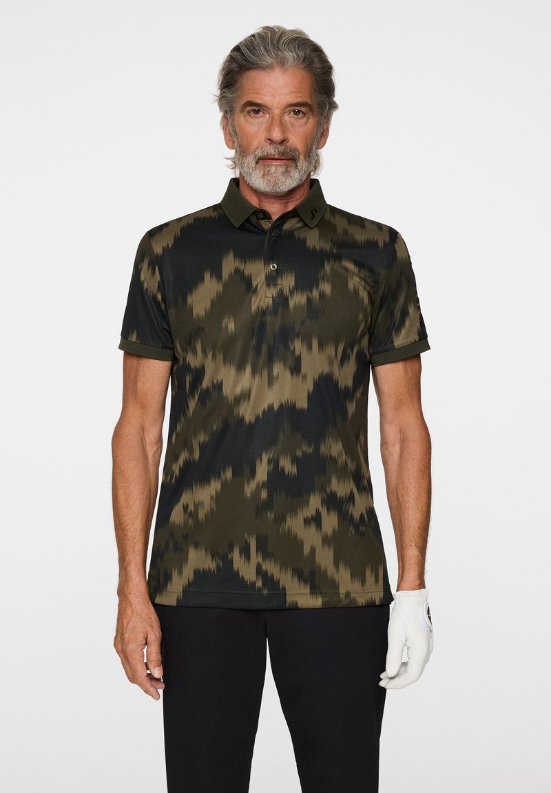 Kortsleeved polo tröja i mörkgrön och svart, med ett zigzag-mönster, knappslå och krage, kombinerad med svarta byxor och en vit handske.