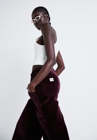 Bordeaux corduroy broek met zijzakken en een 'Lee' label op de achterkant. Model draagt een witte geribde tanktop en lichte zonnebril.
