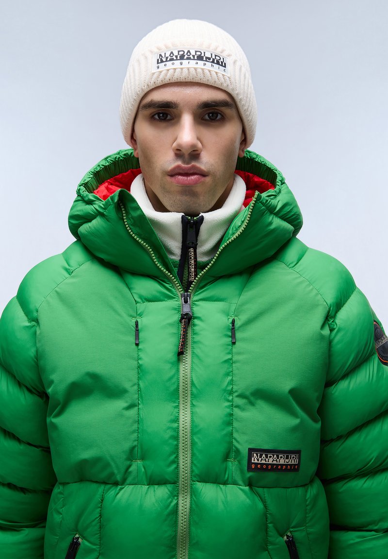 Giacca gonfiabile verde con colletto alto, caratterizzata da una chiusura frontale con zip, hardware nero e un patch logo ricamato sul lato sinistro del petto. Berretto di maglia bianco.