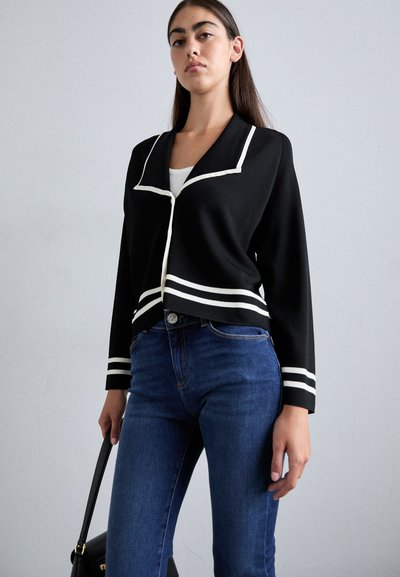 Svart kort jacka med vit kant och ränder, långa ärmar och krage. I kombination med blåjeans och en svart handväska.