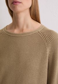 Beige gebreide trui met een gestructureerd patroon, voorzien van een ronde halslijn en raglanmouwen, die een lichte en ademende stof toont.