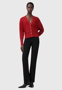 Cardigan rosso lavorato a maglia con scollo a V profondo e chiusure con bottoni. Indossato sopra pantaloni neri svasati, abbinato a scarpe nere a punta.