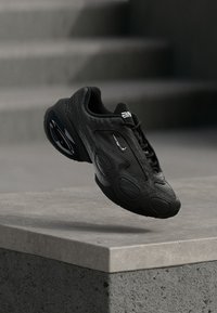 Μαύρο αθλητικό παπούτσι με υφασμάτινο επάνω μέρος με υφή, στρογγυλεμένο σχήμα, καουτσούκ σόλα και έντονες κυκλικές λεπτομέρειες. Διαθέτει λευκό λογότυπο κοντά στα κορδόνια.