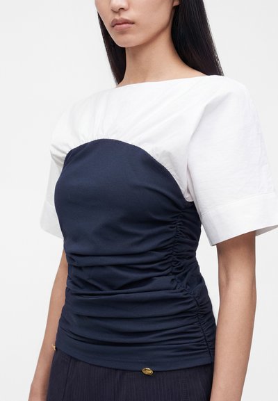 Femme portant un haut blanc à manches courtes superposé d’un vêtement style bustier froncé bleu marine sur le torse.