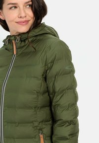 Polstret grøn jakke med hætte, der har en frontlynlås, snøreaccenter og en glat tekstur. Bemærkelsesværdige syninger og imitation læderdetaljer.
