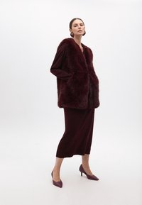 Burgundské umělé kožešinové sako s límcem, vrstvené přes odpovídající šaty, doplněné špičatými fialovými lodičkami a kulatými náušnicemi.