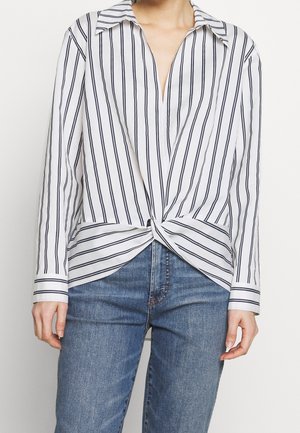 Personne portant une chemise blanche à rayures verticales noires, nouée à la taille, et un jean bleu en denim, visible du cou aux hanches.