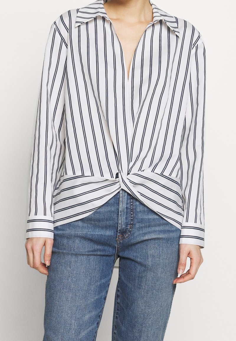 Personne portant une chemise blanche à rayures verticales noires, nouée à la taille, et un jean bleu en denim, visible du cou aux hanches.