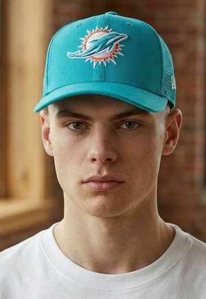 Junger Mann trägt eine türkisfarbene Baseballkappe mit dem Miami Dolphins-Logo und ein weißes T-Shirt, schaut nach vorne mit neutralem Gesichtsausdruck, drinnen.