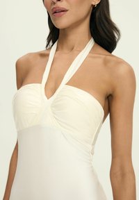 Sukienka na halter w miękkim, jasnym kremowym materiale z marszczoną górą i gładką, dopasowaną spódnicą. Posiada lekki połysk i eleganckie detale drapowania.