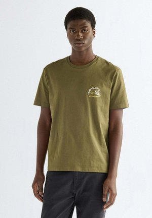 Giovane uomo che indossa una t-shirt verde oliva con il logo "Denim Club Wrangler" sul petto sinistro, abbinata a pantaloni neri, in piedi davanti a uno sfondo bianco.