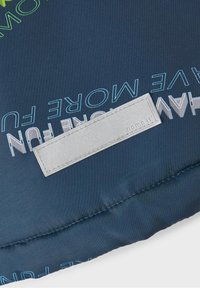 Modrá polstrovaná látka s embosovaným textem "MĚJTE VÍCE ZÁBAVY" a stříbrnou nálepkou s nápisem "nome.it", která je přišitá. Hladký povrch a matný finiš.