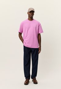 Ροζ βαμβακερό μπλουζάκι με κοντά μανίκια, άνετη εφαρμογή, συνδυασμένο με σκούρο μπλε τζιν παντελόνι και καφέ παπούτσια, ολοκληρωμένο με μπεζ καπέλο.