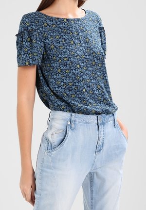Kvinde iført en blå blomstret kortærmet bluse stoppet ned i lyseblå højtsiddende jeans, stående med den ene hånd i lommen.