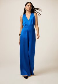 Mono azul real con un profundo escote en V, piernas anchas y cintura ajustada. Hecho de una tela suave y brillante. Combinado con zapatos negros de punta.