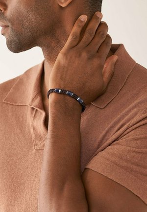 Braccialetto intrecciato nero con perline rettangolari blu, indossato al polso di una persona con pelle scura, su uno sfondo neutro.