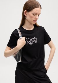 T-shirt en coton noir avec un logo blanc "Calvin Klein", manches courtes et coupe décontractée. Associé à un sac transparent.