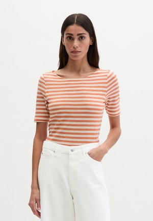 Femme aux longs cheveux foncés portant un t-shirt ajusté à rayures orange et blanches à manches courtes et un pantalon blanc taille haute, debout avec une main dans la poche.