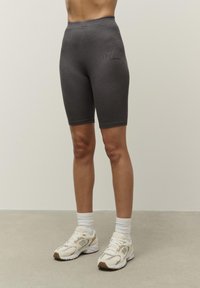 Sivé cyklistické šortky s bezšvovým dizajnom, vysokým elastickým pásom a decentným logom. Nosia sa s bielymi ponožkami po členky a béžovými bežeckými topánkami.