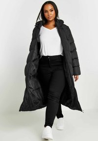 Svart vadderad långjacka med huva och dragkedja, med en quiltad design. Bärs över en vit V-ringad tröja och svarta byxor. Vita sneakers.