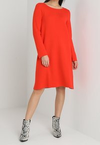 Rotes Langarmkleid mit Rundhalsausschnitt, das einen lockeren Schnitt und einen oberhalb des Knies endenden Saum aufweist. Kombiniert mit schwarz-weißen Stiefeletten im Schlangenmuster.