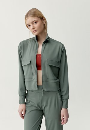 Mujer joven con chaqueta y pantalones de color verde salvia con cremallera y un sujetador deportivo rojo, de pie frente a un fondo claro y liso.