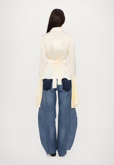 Bluză albă texturată, cu mâneci lungi și evazate, și talie legată, purtată cu blugi supradimensionați din denim albastru, cu buzunare aplicate.