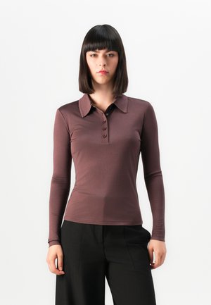 Woman with straight dark hair wearing a fitted long-sleeve brown polo shirt and black wide-leg pants against a plain white background.