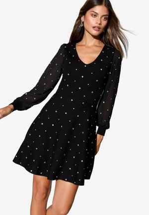 Frau in einem schwarzen Kleid mit durchsichtigen langen Ärmeln und silbernen Polka-Dots, posiert mit fließenden Haaren und schaut zur Seite.