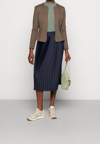 Femme portant une veste marron, un haut côtelé vert, une jupe midi plissée navy, des baskets beiges, tenant un sac à main vert clair sur un fond gris.