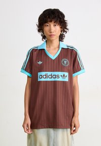 Hnedý pruhovaný dres s svetlomodrým golierom a akcentmi. Obsahuje logo "adidas" a kruhový emblém na hrudi. Krátke rukávy.