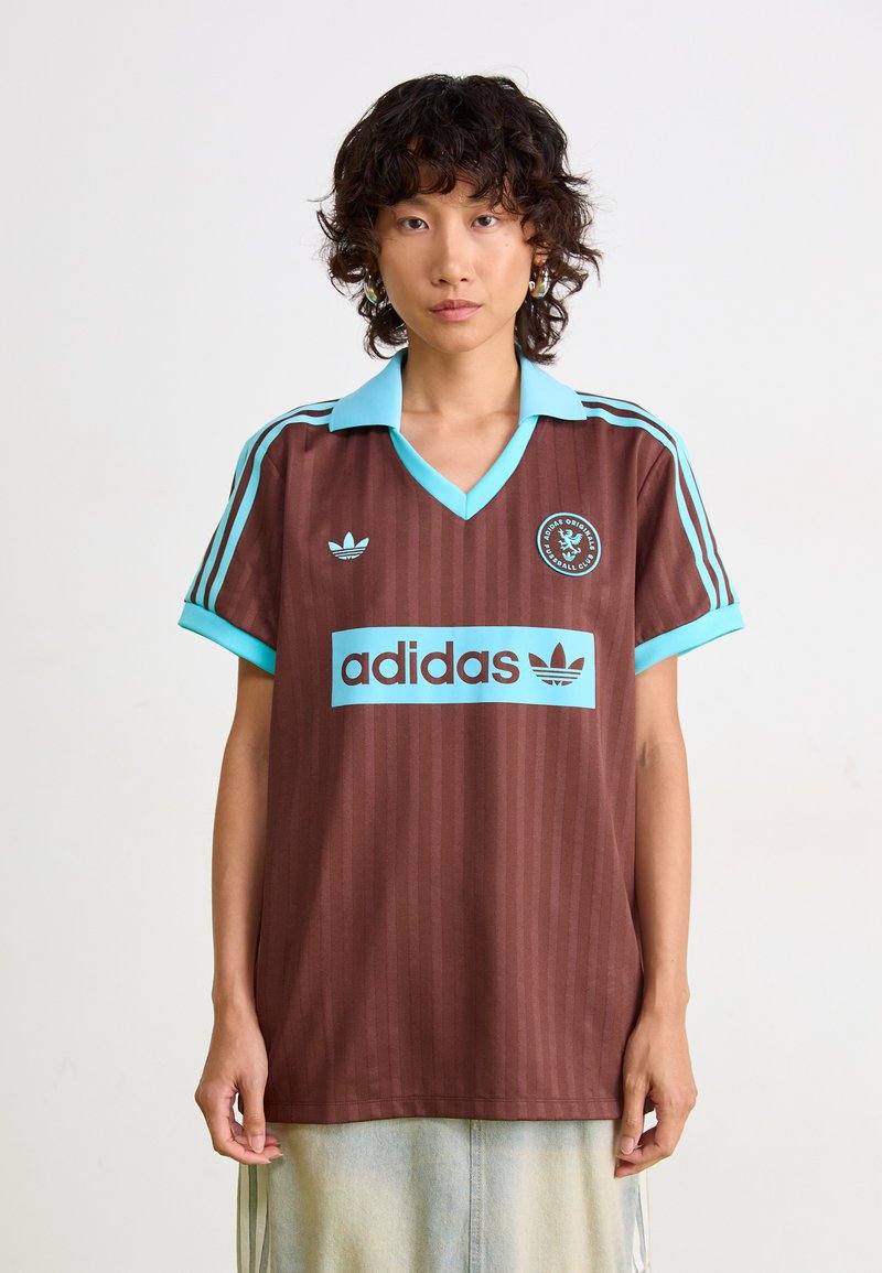 Hnedý pruhovaný dres s svetlomodrým golierom a akcentmi. Obsahuje logo "adidas" a kruhový emblém na hrudi. Krátke rukávy.