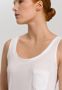 Top in cotone bianco senza maniche con scollo rotondo, spalline sottili e una piccola tasca sul petto. Tessuto morbido e leggero con una texture liscia.