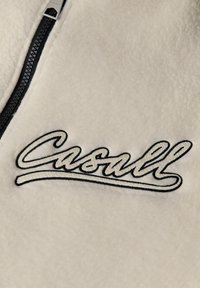Krämfärgad fleeceklädsel med dragkedja, med en svart broderad "Casall"-logotyp. Slät textur och kontrasterande svarta detaljer längs dragkedjan.