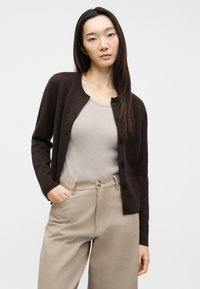 Γυναίκα με μακριά σκούρα μαλλιά, φοράει καφέ σκούρη ζακέτα με κουμπιά πάνω από μπεζ ριμπάτο τοπ και μπεζ παντελόνι ψηλόμεσο, με το χέρι στην τσέπη.