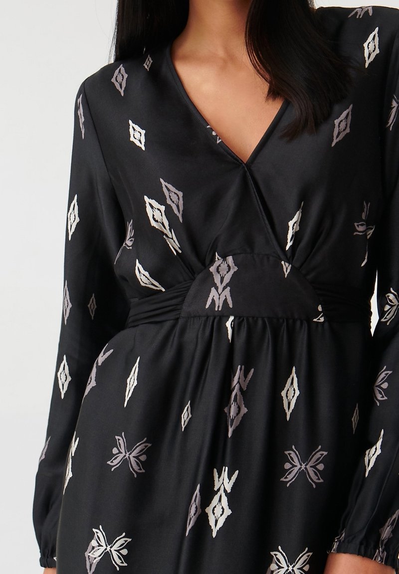 Femme portant une robe portefeuille noire à manches longues avec des motifs losanges et papillons blancs et gris, montrant le haut du torse et le cou.