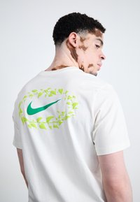 Hvid bomulds t-shirt med et grønt Nike-logo omgivet af stiliserede bladmønstre på ryggen. Klassisk crew neck og korte ærmer.