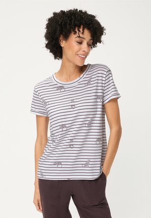 Frau trägt ein weißes und braun gestreiftes Kurzarmshirt mit kleinen Leopardenillustrationen, kombiniert mit dunklen Hosen, steht mit einer Hand in der Tasche.
