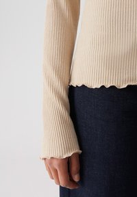 Beige gerippter Langarmoberteil mit ausgestellt Ärmeln und gewelltem Saum, getragen mit dunkelblauen Jeans.