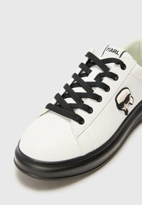 Witte leren sneaker met zwarte veters, een zwarte rubberen zool en een zwart-wit logo op de zijkant. Gladde textuur en een ronde teenvorm.