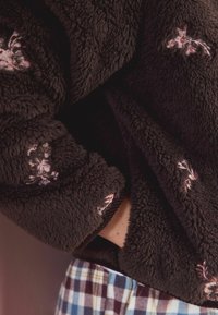 Minkštas, tamsiai rudas fliso megztinis su rožinėmis gėlių siuvinėjimais. Turime rūbų su elastingu kraštu ir laisvu siluetu, derinamas su margais pižamų kelnėmis.