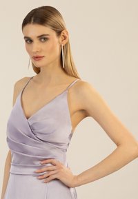 Lilafarbene Satin-Kleid mit tiefem V-Ausschnitt, Drapierung und Spaghetti-Trägern. Das Model trägt lange silberne Ohrringe, das Haar ist glatt frisiert.
